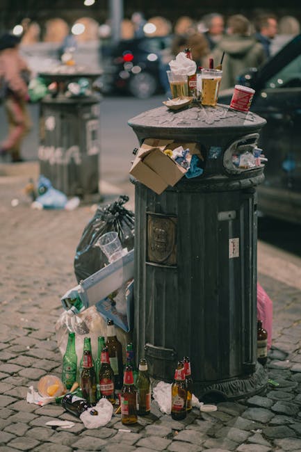 A black cylindrical rubbish bin with a domed lid, situated on a cobblestone surface in an urban outdoor setting, shows various discarded items on top and around it. The top of the bin holds several empty plastic cups and glasses, some containing liquid or foam, with a few straws, along with a crumpled paper cup. A small cardboard box is perched on the rim, and underneath, the side of the bin has a bag of trash partially hanging out, alongside a collection of glass bottles, some with labels visible, and other waste items like paper and plastic debris scattered at the base. In the background, out of focus, are pedestrians walking, parked cars, and a grey metal waste container, indicating a busy city street environment. The scene reflects typical street-level litter near public seating or dining areas, illustrating the importance of efficient rubbish removal services provided by companies like House Clearance Marylebone to maintain cleanliness and manage waste effectively.
