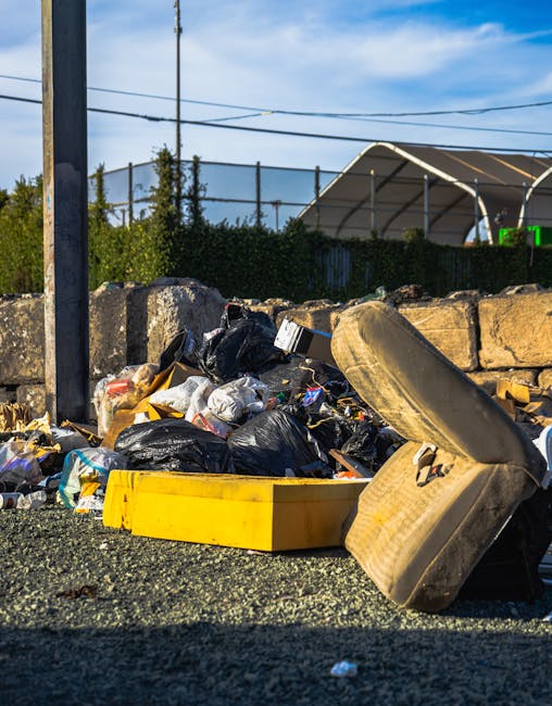 A black cylindrical rubbish bin with a domed lid, situated on a cobblestone surface in an urban outdoor setting, shows various discarded items on top and around it. The top of the bin holds several empty plastic cups and glasses, some containing liquid or foam, with a few straws, along with a crumpled paper cup. A small cardboard box is perched on the rim, and underneath, the side of the bin has a bag of trash partially hanging out, alongside a collection of glass bottles, some with labels visible, and other waste items like paper and plastic debris scattered at the base. In the background, out of focus, are pedestrians walking, parked cars, and a grey metal waste container, indicating a busy city street environment. The scene reflects typical street-level litter near public seating or dining areas, illustrating the importance of efficient rubbish removal services provided by companies like House Clearance Marylebone to maintain cleanliness and manage waste effectively.