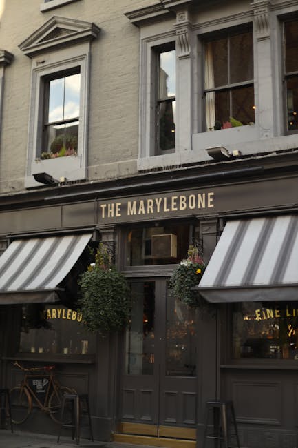 The exterior facade of a building featuring a pub named 'The Marylebone', with a dark grey painted front and gold lettering displaying the pub's name. Two large black and white striped canvas awnings extend over the windows on either side of the entrance, which has a central glass door with a gold handle. Above the door, there is a window with a window box filled with small green plants. The building has three upper-story windows with decorative white trim, and potted plants can be seen inside through the glass panes. The environment appears to be part of a street scene with natural daylight illuminating the facade, giving a clear view of the pub’s exterior details which are relevant for an illustration of premises involved in privately managed waste disposal and rubbish collection services, consistent with professional waste removal contexts in urban areas.