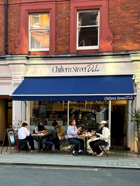 Four people sit outside Chiltern Street Deli, a small café with a white façade and a dark blue awning displaying its name. The group includes a man in a light grey jacket, a man in a white cap and dark trousers, and two women, one wearing a black top and the other a tan sweater. They are seated at tables with black and wooden chairs, enjoying drinks and conversation on the sidewalk in front of the deli. The café's interior is partially visible through large glass windows, revealing shelves stocked with various items and a brightly lit space. Above the storefront, two white-framed windows with clear glass are set into a red-brick building, which has decorative stonework and circular motifs on either side of the windows. The scene suggests an urban setting with natural daylight illuminating the outdoor seating, and the overall atmosphere appears casual and inviting, aligned with services related to private rubbish handling and independent collection for commercial establishments like cafes.