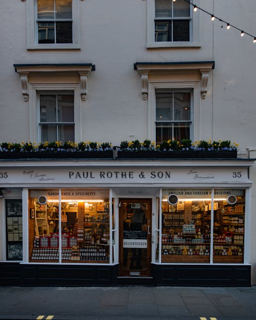 The exterior facade of a building featuring a pub named 'The Marylebone', with a dark grey painted front and gold lettering displaying the pub's name. Two large black and white striped canvas awnings extend over the windows on either side of the entrance, which has a central glass door with a gold handle. Above the door, there is a window with a window box filled with small green plants. The building has three upper-story windows with decorative white trim, and potted plants can be seen inside through the glass panes. The environment appears to be part of a street scene with natural daylight illuminating the facade, giving a clear view of the pub’s exterior details which are relevant for an illustration of premises involved in privately managed waste disposal and rubbish collection services, consistent with professional waste removal contexts in urban areas.