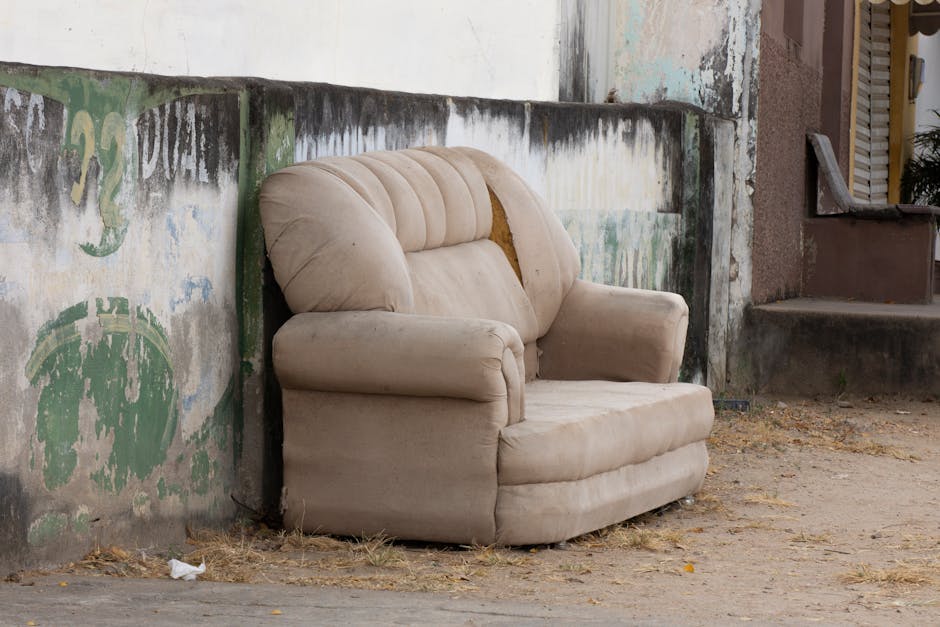 An old, beige upholstered armchair with a worn and slightly sagging fabric, situated outdoors against a weathered concrete wall displaying faded green and black paint remnants and graffiti. The armchair's cushions show signs of age, with visible indentations and minor stains, and the fabric appears frayed in places, especially on the armrests and backrest. The chair's right side is partially obscured by the wall, and it rests on dry, patchy ground with scattered leaves and small debris. Behind the chair, there is a concrete staircase leading to a building with a textured, brown exterior wall and closed window shutters. The scene is illuminated by natural daylight, revealing the textures and muted colors of both the furniture and surrounding environment. This setting, with furniture abandoned on the street, relates to private waste disposal efforts often handled by specialized rubbish removal services, such as those provided by House Clearance Marylebone.