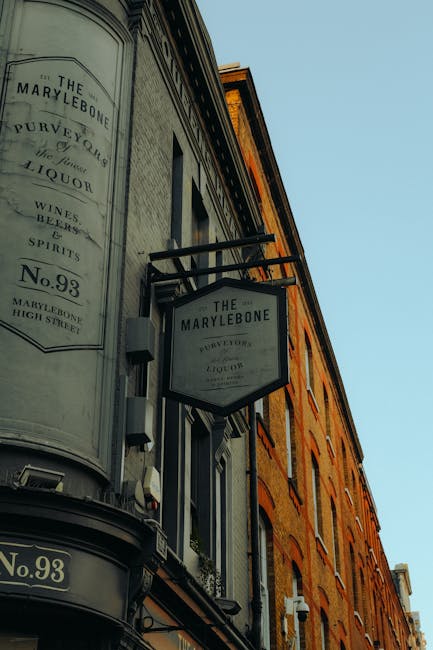 A close-up view of the exterior of a historic-style building with a rounded corner, featuring a large black and white sign hanging from a metal bracket that reads 'The Marylebone,' with smaller text mentioning 'Purveyor of Liquor, Wines, Beers, Spirits' and the address number 'No. 93.' The building's façade is constructed from warm, reddish-brown brick with multiple rectangular windows and decorative brickwork, set against a clear, pale blue sky. The sign and building suggest a traditional pub or liquor shop situated on a high street or lively urban area, indicative of private and independent waste collection needs often associated with local businesses or commercial properties, as managed by services like House Clearance Marylebone. The image emphasizes the classic architectural details and signage typical of Marylebone’s historic commercial streets, relevant for visually supported content on rubbish removal and alternative waste handling solutions for such premises.