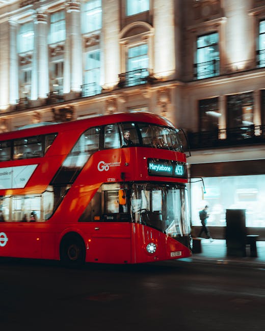 A red double-decker bus operating as a London transport vehicle is moving along a city street in front of historic, multi-story buildings with ornate architectural details, large windows, and balconies. The bus displays the destination 'Marylebone' on its electronic sign, indicating its route towards Marylebone station or area. The bus's exterior is smooth with a glossy finish, reflecting ambient light from the urban environment. The background includes a pedestrian walking on the pavement, while shopfronts and commercial activity are visible through large glass windows, illuminated by interior lighting. The scene captures the dynamic movement typical of city life, with the bus slightly blurred to convey motion, and the surrounding architecture providing a classic London setting. This everyday scene relates to private or independent transport options and alternative ways of navigating busy urban areas, complementing the broader context of rubbish removal and waste management services often integrated into city logistics, as provided by local companies like House Clearance Marylebone.