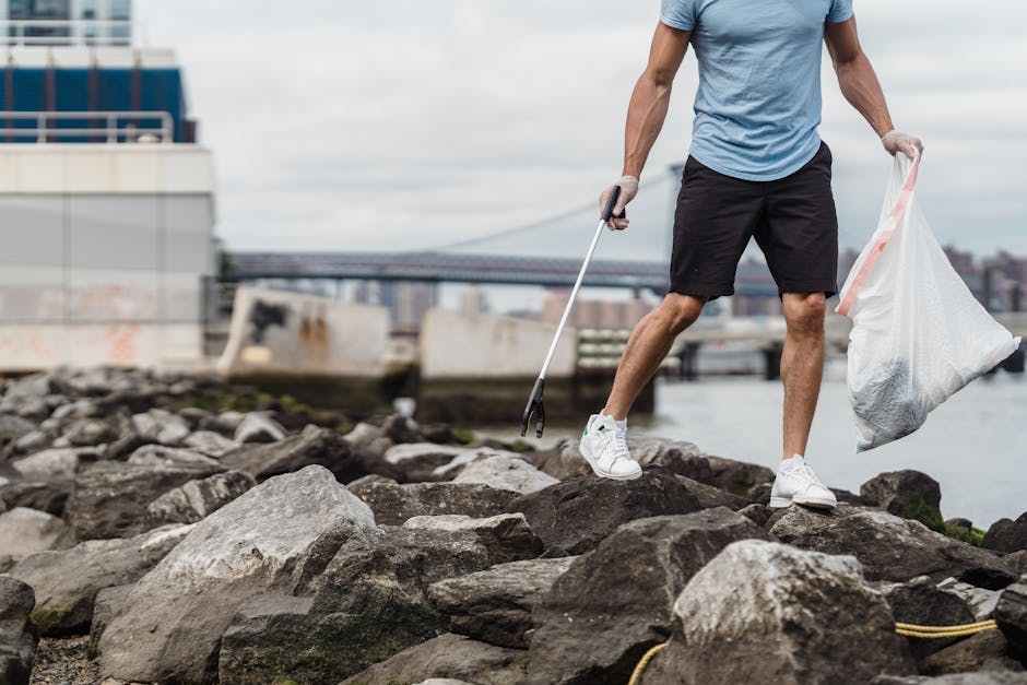 A person wearing a light blue t-shirt, black shorts, and white sneakers stands on a rocky shoreline, holding a large white plastic bag filled with collected rubbish in one hand and using a litter picker in the other to gather debris. The rocks are irregular in shape and grayish-brown in color, with some patches of moss or algae. In the background, there is a cityscape featuring modern buildings, a bridge spanning across the river, and a cloudy sky overhead. The environment appears to be a cleanup effort along a waterfront, emphasizing responsible waste disposal away from typical urban streets. The scene’s natural lighting and outdoor setting highlight the activity of independent rubbish collection, aligning with the principles of private waste handling and environmentally conscious rubbish removal services, such as those offered by House Clearance Marylebone.