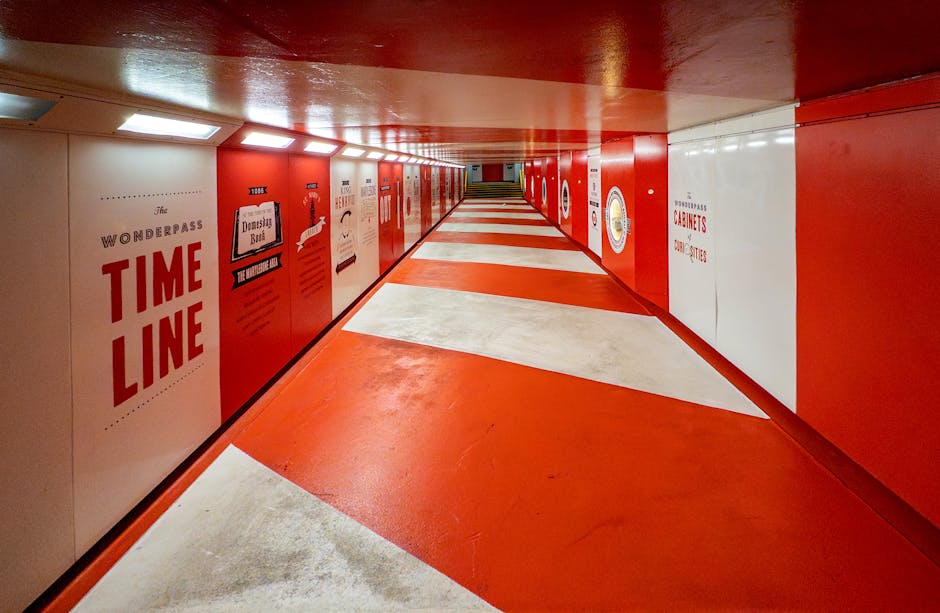 A brightly lit underground corridor with a red ceiling and matching red walls, adorned with various posters and signs, including one reading 'The Wonderpass Time Line' in large red letters. The floor features a pattern of red and white geometric shapes, creating a visually striking pathway that extends into the distance. The corridor's surfaces are smooth, with a polished finish reflecting the light from rectangular ceiling fixtures. On the right side, partially visible, there is a window-like structure or display case set into the wall. The overall environment appears clean and modern, with a vibrant colour scheme that emphasizes the geometric design. This setting resembles a themed public space or an artistic installation, providing a stark contrast to typical rubbish removal environments. House Clearance Marylebone finds relevance in such contexts where alternative waste handling or creative spaces are involved, although the primary subject here remains a structured, decorative corridor rather than a disposal site.
