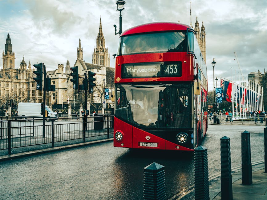 A red double-decker bus is parked on a wet city street with a historic Gothic-style building featuring tall spires and intricate stonework in the background. The bus displays the route number 453 and the destination Marylebone on its electronic sign above the front windshield, which appears slightly fogged. The pavement around the bus is damp, reflecting the overcast sky, and black bollards line the edge of the sidewalk, separating the street from the pedestrian area. To the left, there are several digital traffic lights mounted on black poles, with some showing red signals. In the distance, to the right, there are groups of people walking along the promenade near a row of flagpoles flying various flags. The surrounding environment suggests a busy urban setting, characteristic of areas with regular public transportation and organised city infrastructure, consistent with locations where private rubbish collection services like House Clearance Marylebone might operate for on-site disposal or clearance of waste alongside public infrastructure.