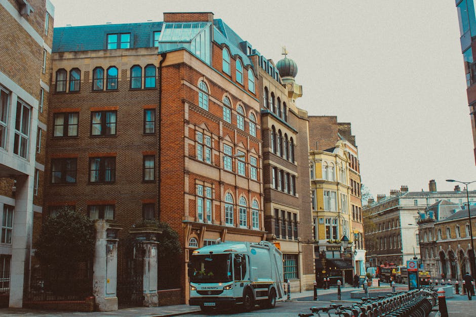 A street scene in an urban area showing a variety of multi-storey buildings with different architectural styles. The prominent building in the center is constructed with red brick and beige stone accents, featuring arched and rectangular windows, a blue-green sloped roof, and small decorative elements. To the right, there is a cream-colored building with ornate detailing, bay windows, and a cornice. In the foreground, a street-side utility vehicle with a white and blue body is parked close to a sidewalk that has metal bike racks and bollards, possibly indicating a designated area for waste collection or loading activities. The environment is lit by natural daylight, casting soft shadows on the pavement and buildings, suggesting a clear day. The setting appears to relate to private waste handling, as indicated by the presence of a collection vehicle and the urban surroundings, aligning with local rubbish removal services offered by House Clearance Marylebone in a central London location.