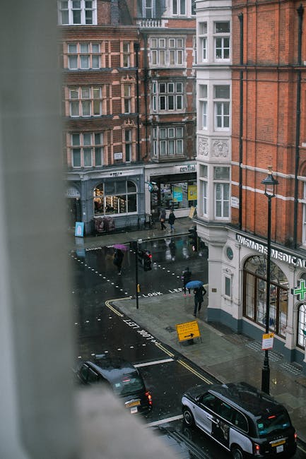A view from an upper window overlooking a wet city street in Marylebone, showing pedestrians walking with umbrellas on a rainy day. The street is lined with historic, red-brick and white stone buildings with large bay windows. A small commercial unit with various signs and storefronts is visible at street level. Parked cars are along the curb, and there is a yellow temporary road sign indicating a temporary restriction. The scene captures the typical urban environment where private waste collection services, such as those provided by House Clearance Marylebone, might operate for rubbish removal or house clearance projects, particularly in shared or commercial properties in the area.