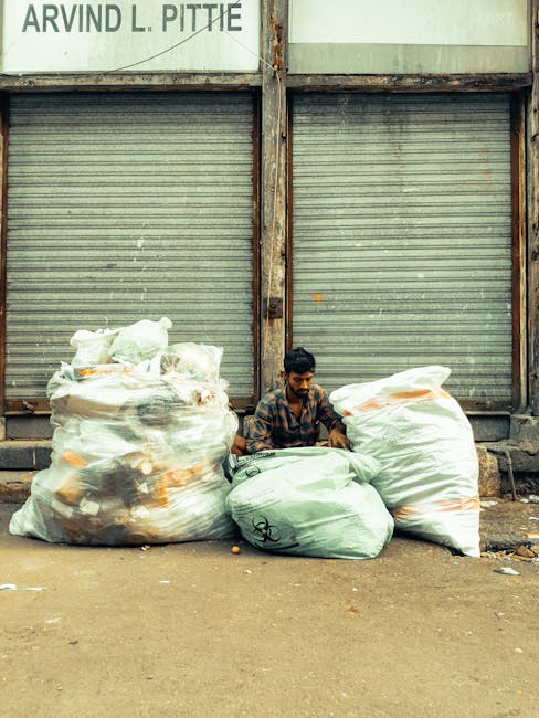 A young man with dark hair and a beard, wearing a checkered shirt, is seated on the ground in front of a corrugated metal warehouse door that is weathered and partially rusted. He is surrounded by large, tightly bundled plastic rubbish bags containing mixed waste, with some bags appearing transparent or semi-opaque, revealing waste materials inside. The bags on his left are mainly white, while the ones on his right are light green, all resting directly on a dusty, uneven pavement surface. Above the doors, there is a partially visible sign with black text, including the name 'ARVIND L. PITTIE.' The scene appears to be a typical urban waste collection or disposal area, with the man possibly involved in rubbish sorting or collection as part of an independent waste handling activity, reflecting the context of private rubbish removal services or street-level waste management. The environment suggests an outdoor setting, with no other objects, vehicles, or people visible in the background, emphasizing the focus on waste bags and the individual's activity amidst an industrial backdrop.