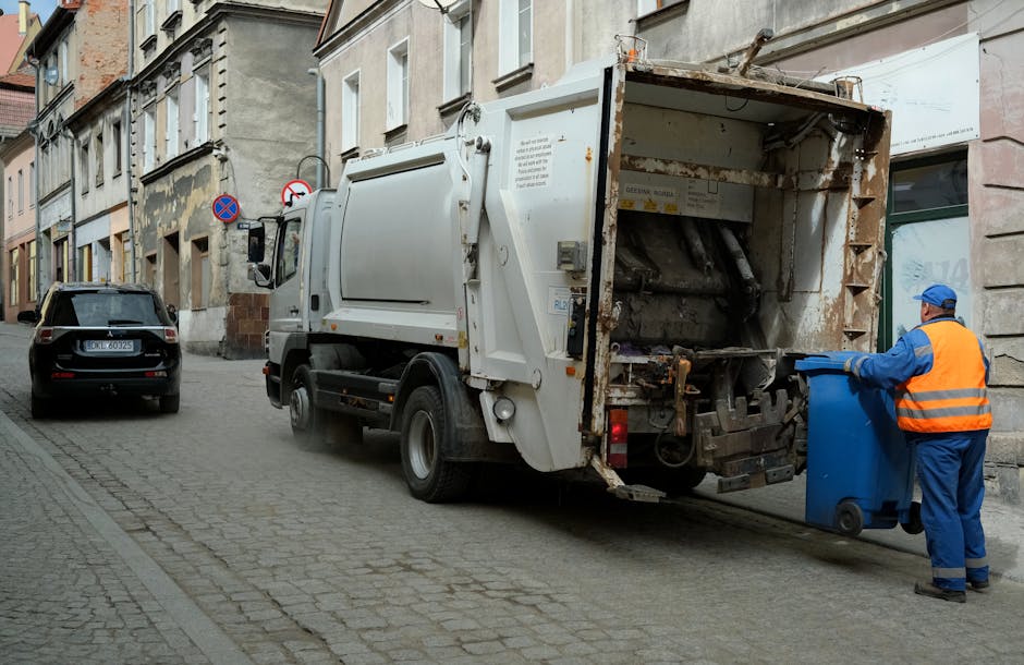 A waste collection vehicle parked on a narrow, urban cobblestone street with old, multi-story buildings on either side. The truck is white with rust and dirt visible on its rear, which is open and equipped with mechanical lifting components for waste handling. A sanitation worker wearing a blue uniform and a bright orange high-visibility vest is standing beside the truck, holding a blue wheelie bin in front of the open rear compartment, ready for waste disposal. The street features parked cars, including a black vehicle directly behind the waste truck, and a street sign visible on a building wall indicating parking restrictions. The scene is captured in natural daylight, with an overall neutral tone, emphasizing the routine activity of rubbish collection, possibly as part of private waste management or alternative collection services like those offered by houseclearancemarylebone.co.uk, serving local waste removal needs without municipal involvement.